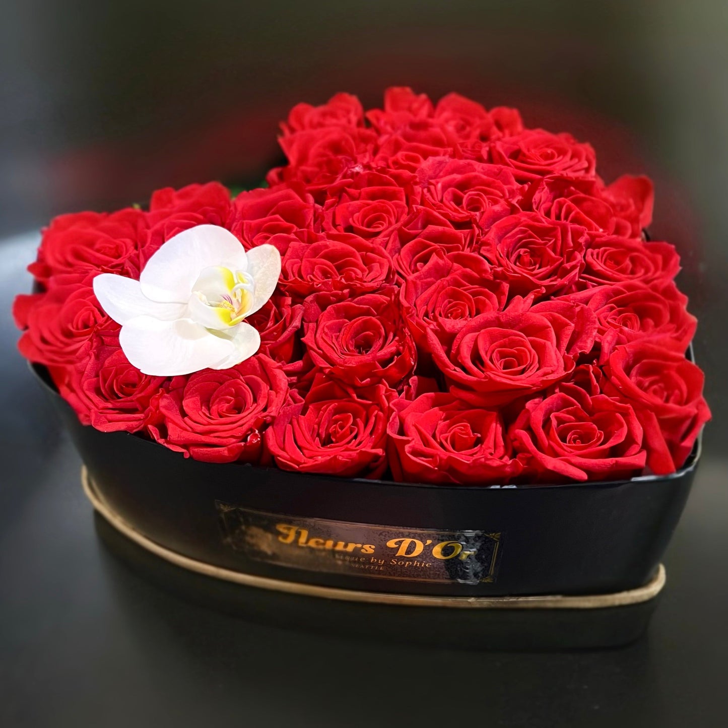 A heart-shaped box filled with red roses, with a single white orchid on top.